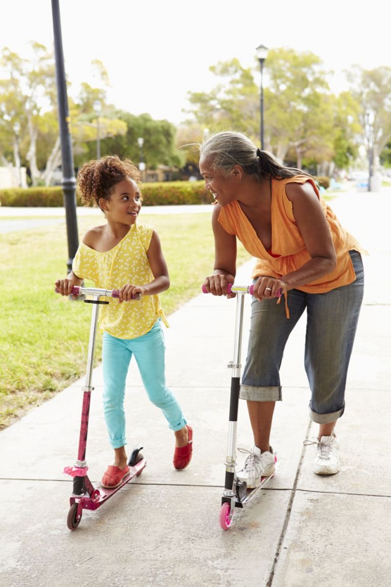A mother and her daughter ride scooters outdoors together