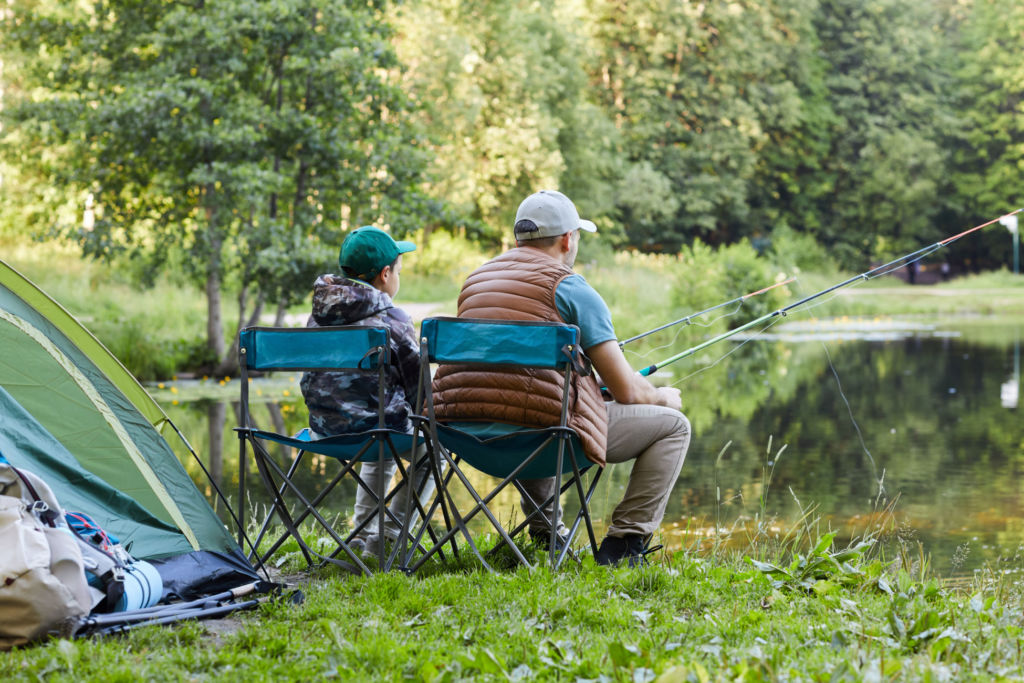 Back view portrait of loving father and son fishing by lake together during camping trip in nature, copy space