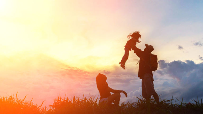 The happy family of three people, mother, father and child in front of a sunset sky.