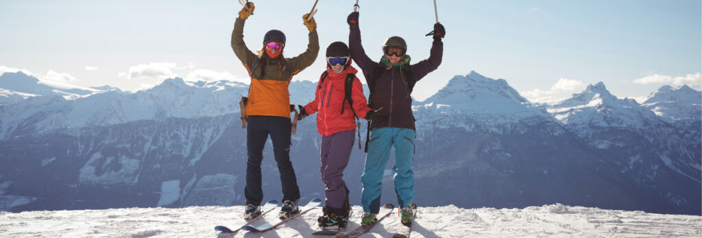 three skiers stand in joyful pose at the top of a mountain