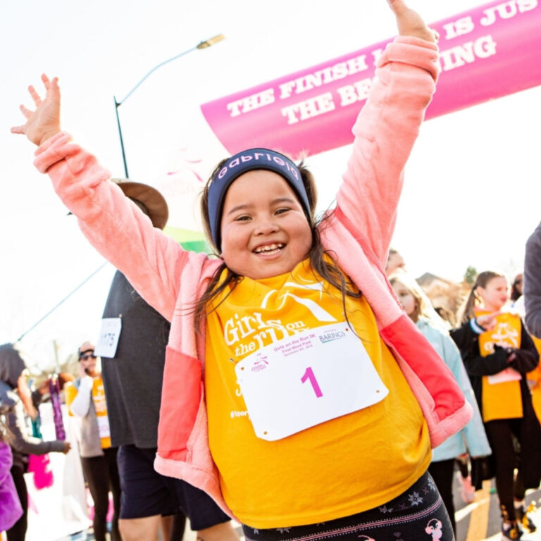 a young girl raises her arms in triumph at the end of the Girls on the Run 5K