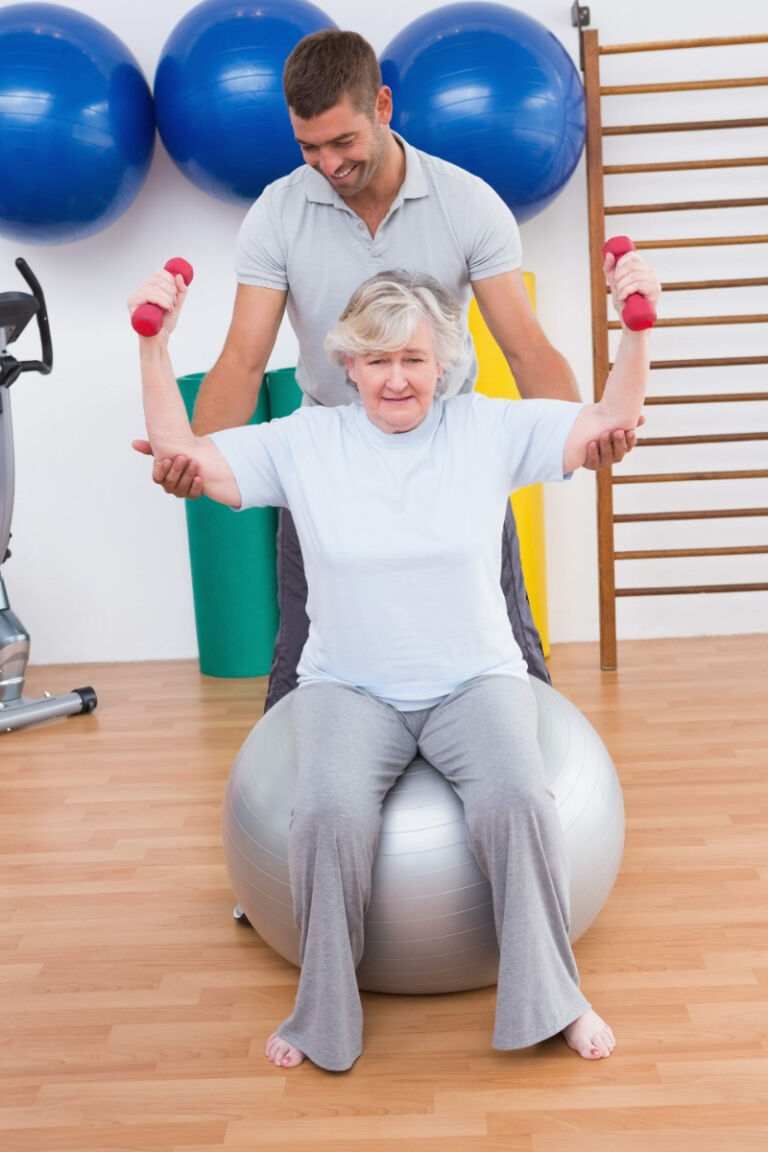 older woman sits on exercise ball lifting dumbbells with PT helping her
