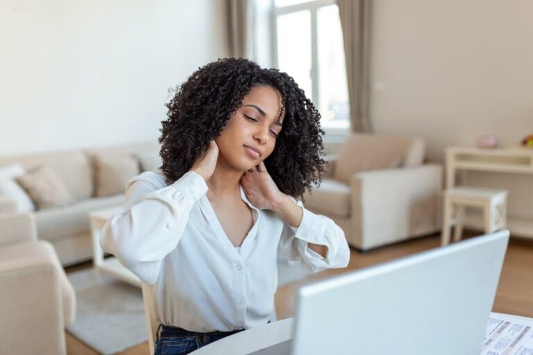 a young woman rubs her neck from pain while sitting working at a laptop computer in a poor ergonomic set up