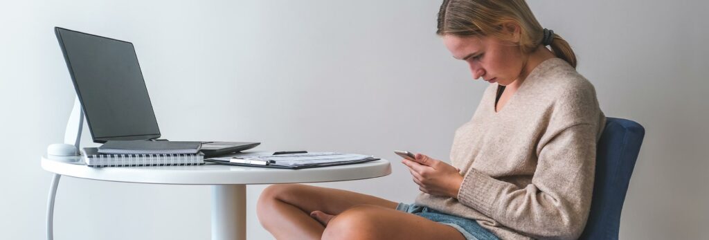 a young woman sits hunched over her cell phone, in a posture that leads to tech neck