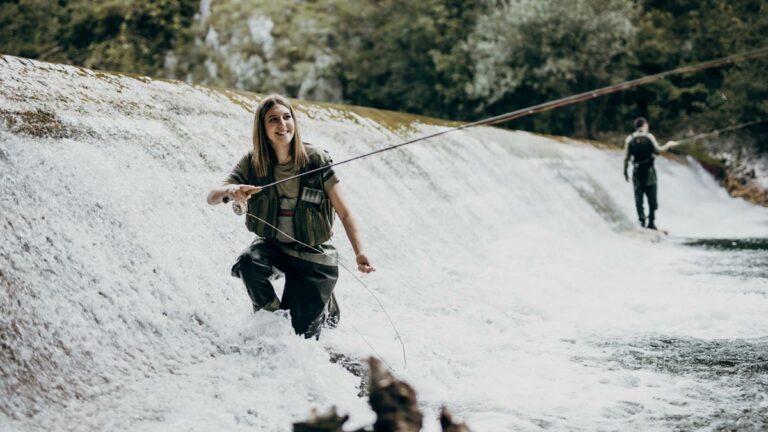 a woman stands in a waterfall of rushing water fly fishing
